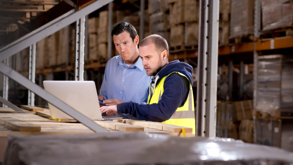 Two warehouse workers look at their TMS, or transportation management system, on a laptop