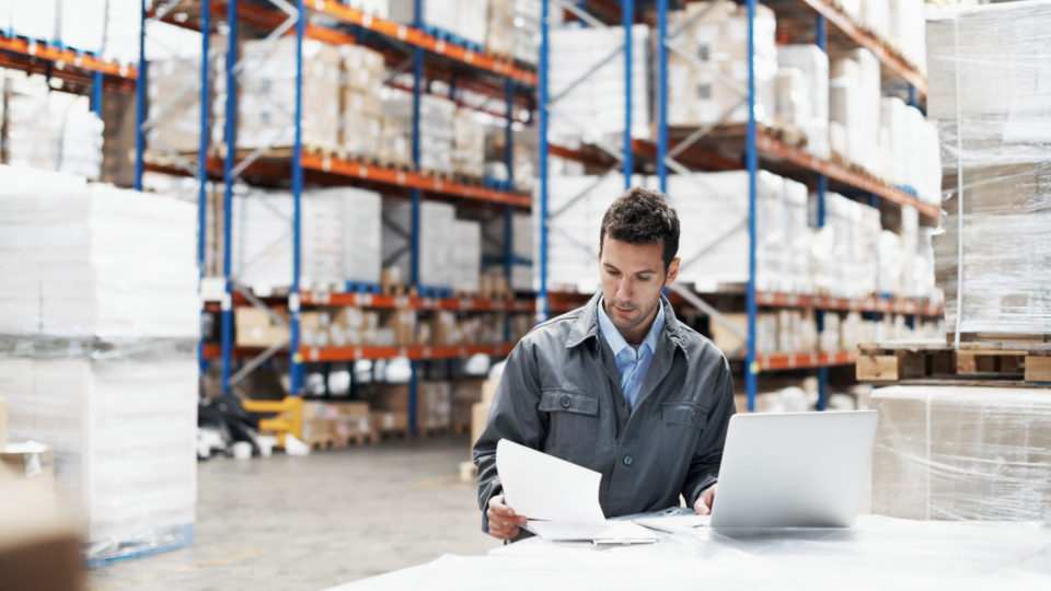 A warehouse worker looks at a laptop and papers