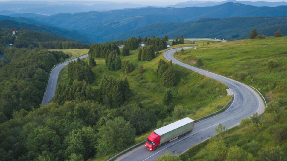 A semi-truck drives on a winding road with mountains in the background