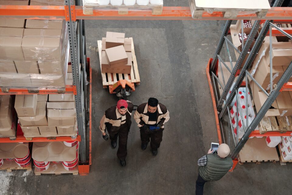 two men in a warehouse pulling a pallet jack walking past another man on a tablet.